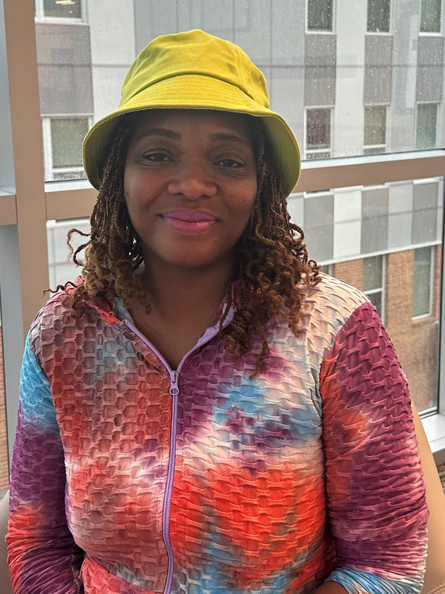 Image of Anita - a black woman with shoulder-length hair, wearing a bright tye-dye zip-up shirt and a yellow bucket hat